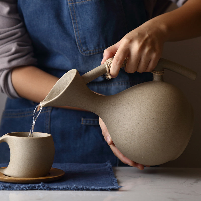 Woman in denim apron pouring water from pottery kettle into ceramic cup with wooden saucer