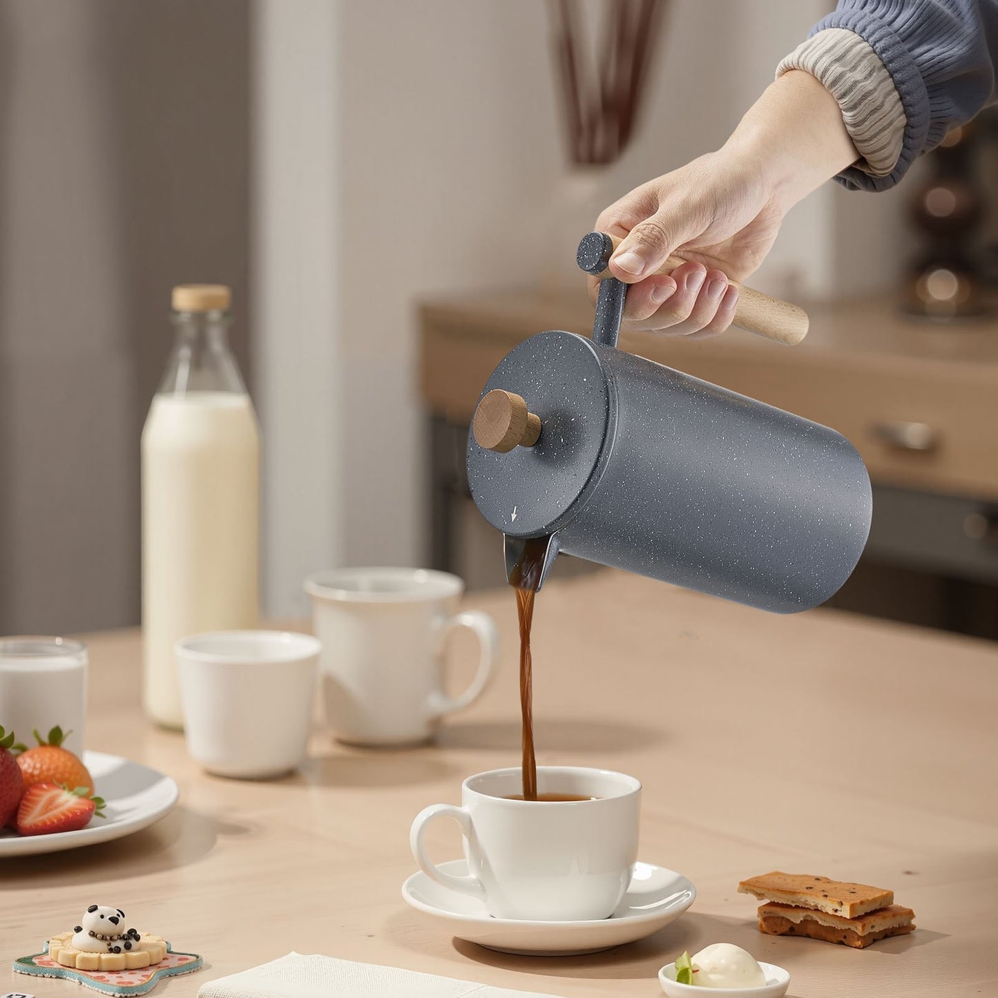 Stainless steel French press pouring coffee into white cup on kitchen table lifestyle