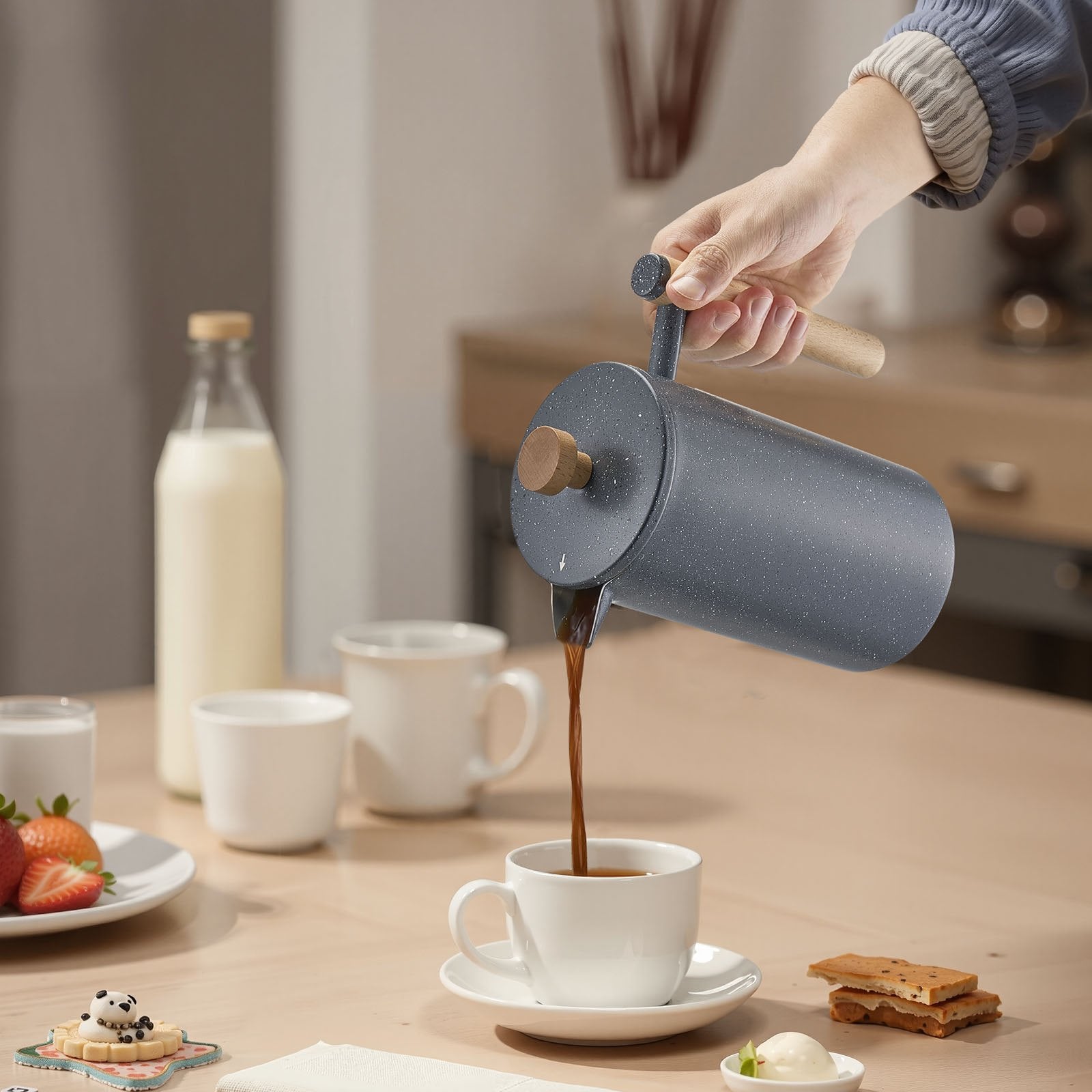Stainless steel French press pouring coffee into white cup on kitchen table lifestyle