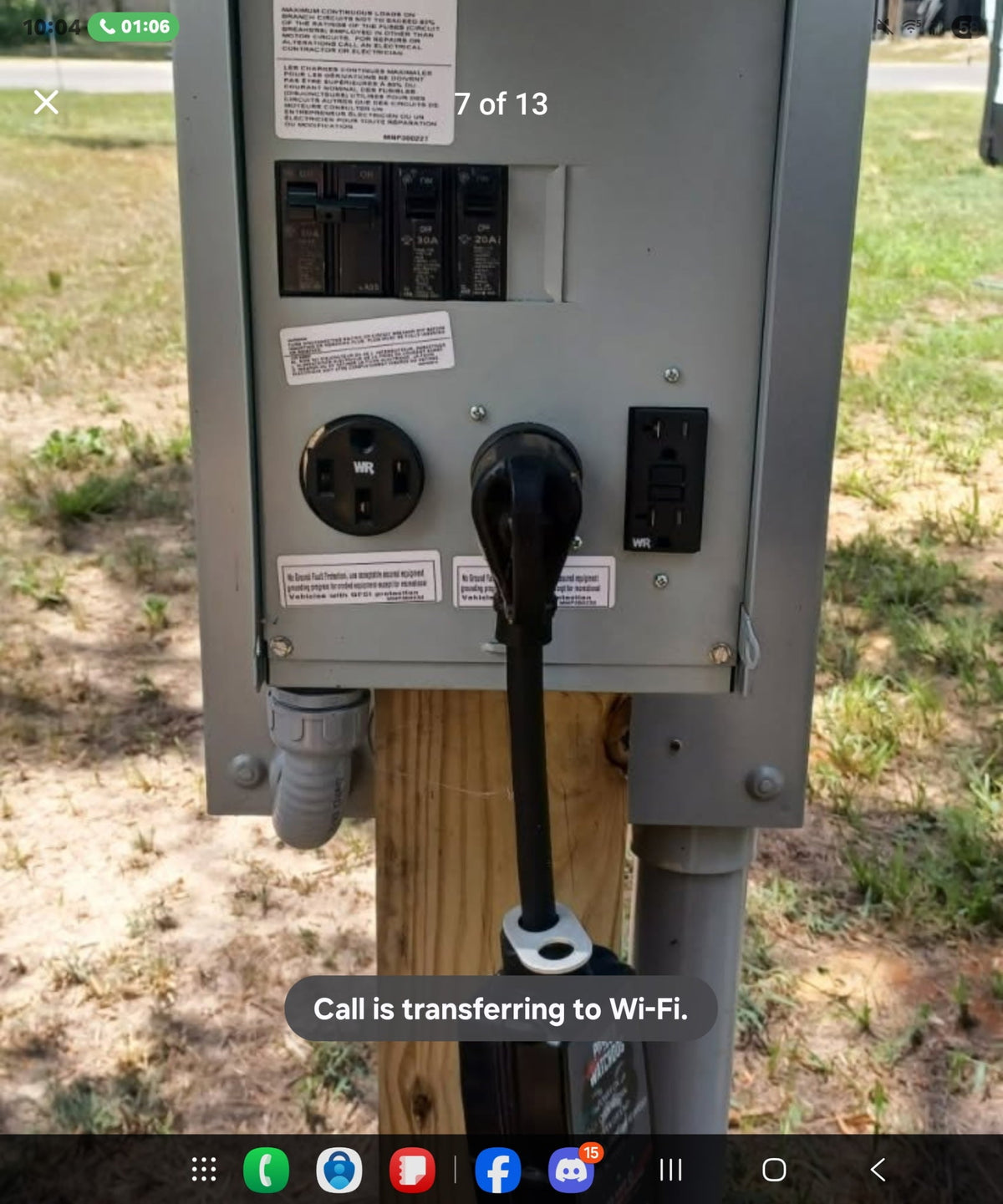 A gray electrical utility panel mounted on a wooden post outdoors, with multiple outlets, circuit breakers, and a black power cord plugged in. A smartphone interface and transfer message are visible on the screen.
