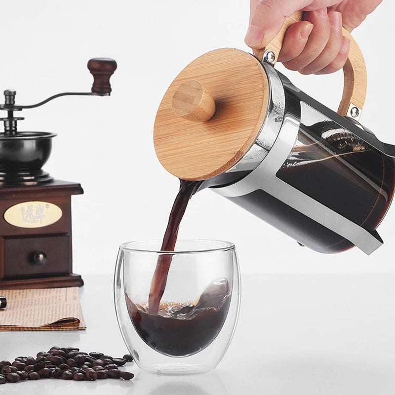 Person pouring coffee from a wooden-lidded French press into a glass, with a wooden grinder and coffee beans in the background.