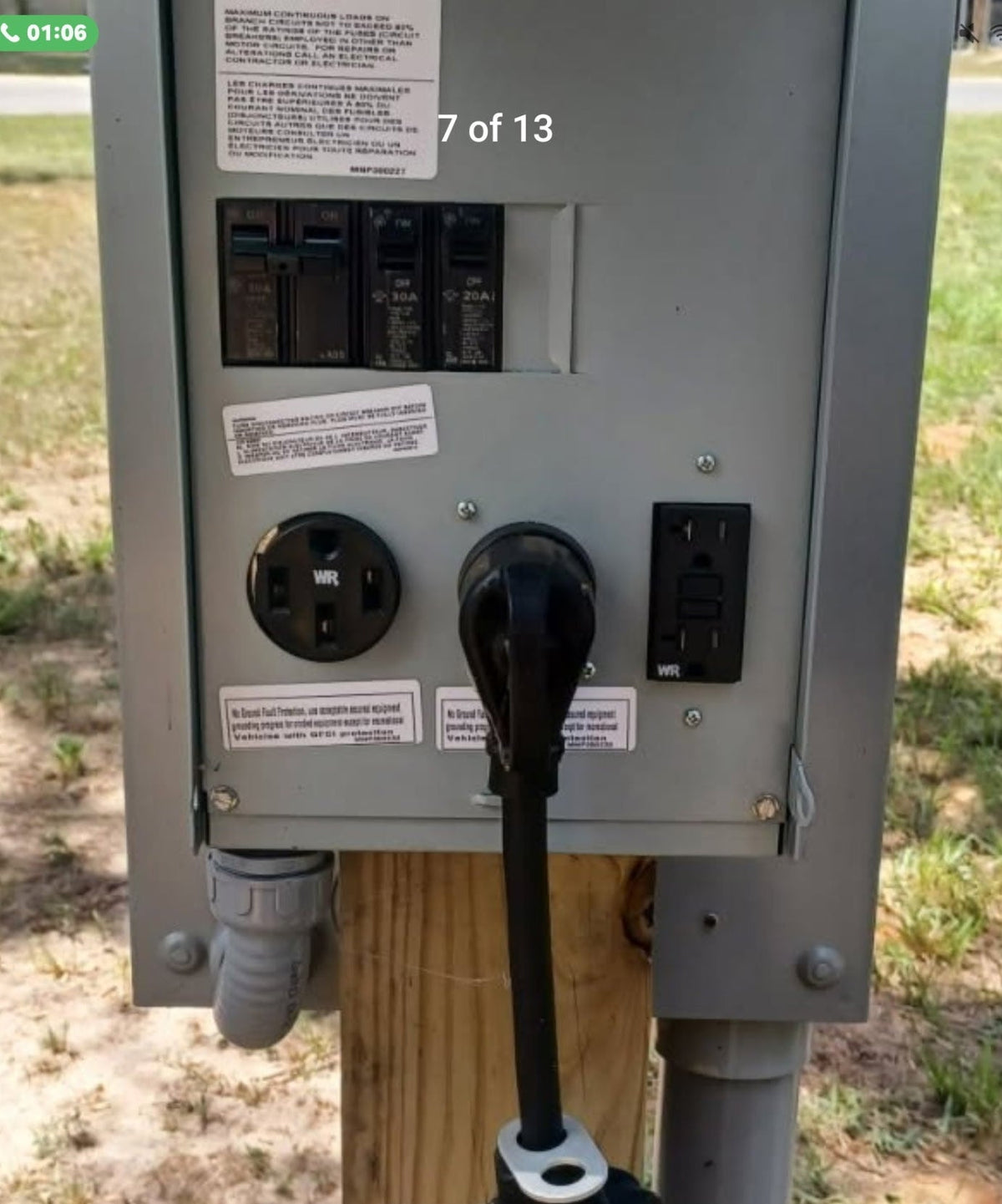 A gray electrical utility panel mounted on a wooden post outdoors, with multiple outlets, circuit breakers, and a black power cord plugged in. A smartphone interface and transfer message are visible on the screen.