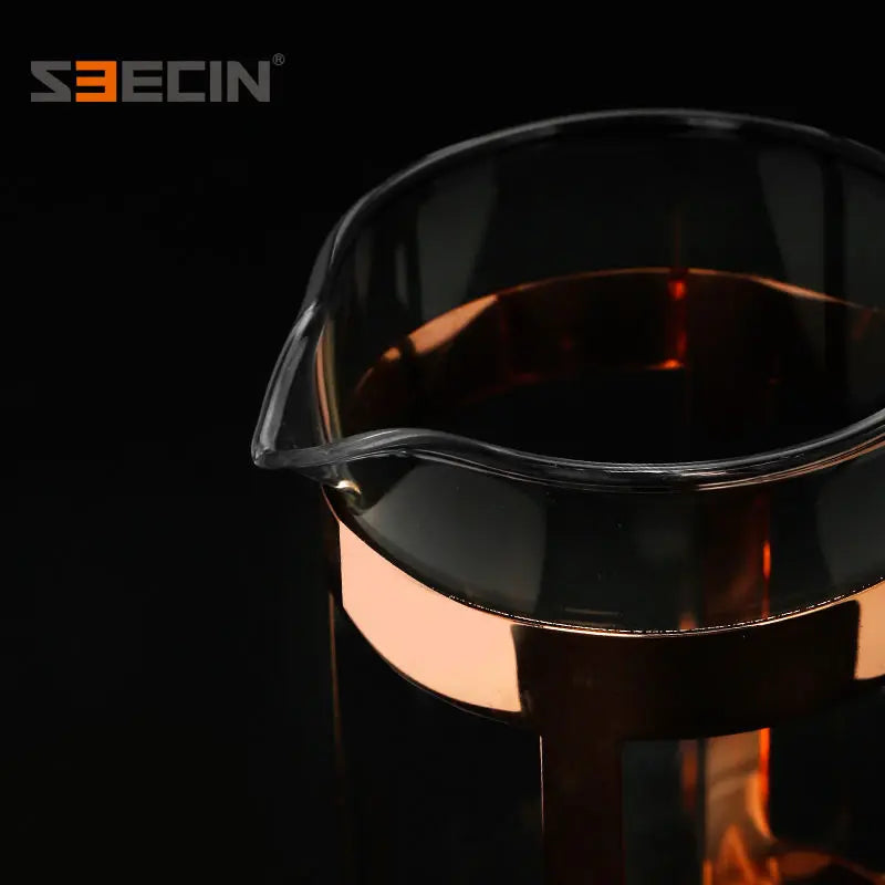 A close-up of a transparent glass pitcher or decanter with a metallic rose-gold or copper-toned band and base, shown against a black background. The S3ECIN brand logo is visible in the top left corner.