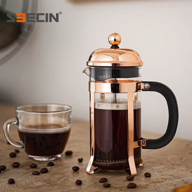 A copper-finished French press coffee maker with a black handle and glass carafe filled with black coffee, shown on a tabletop beside a glass cup of coffee with scattered coffee beans. The S3ECIN brand logo is visible in the top left corner.
