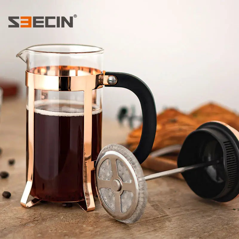 A glass French press coffee maker with a rose gold metal frame and black handle, filled with dark coffee, shown on a wooden surface with the plunger lid placed beside it. The S3ECIN brand logo is visible in the top left corner.