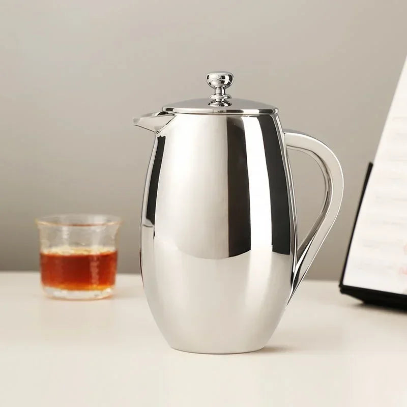 Stainless steel coffee press on a table with a glass of liquid and laptop in the background