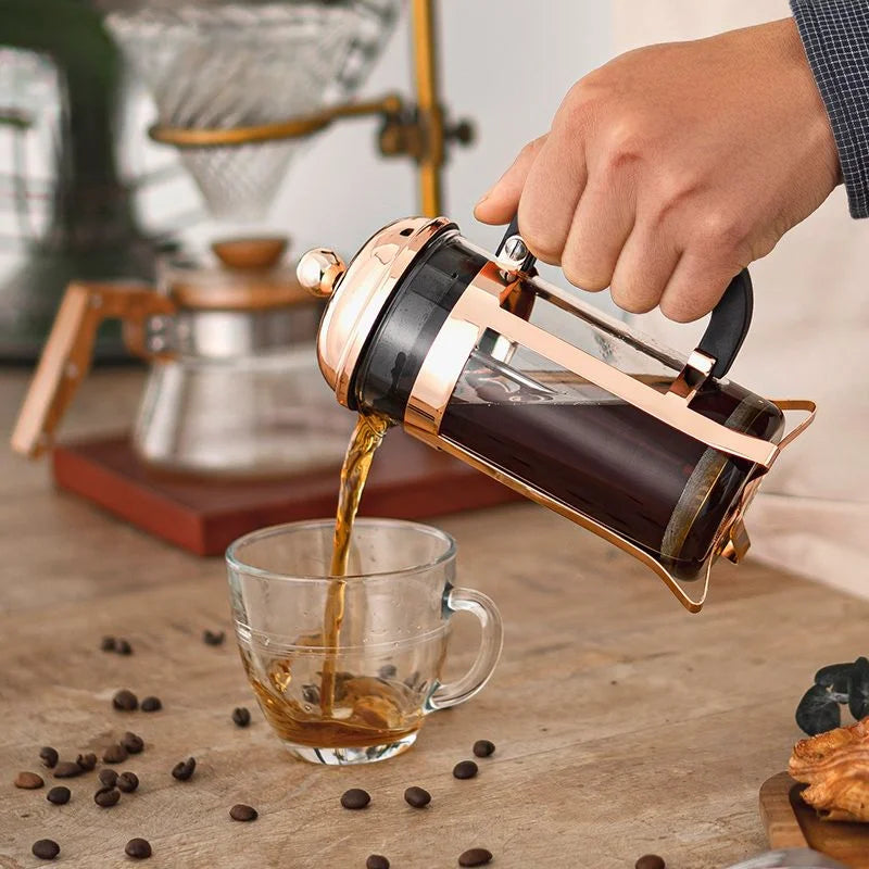 A hand pouring brewed coffee from a copper-and-glass French press into a clear glass cup on a wooden table, with coffee beans scattered around and a coffee maker in the background.