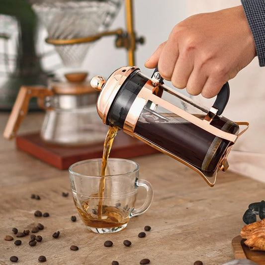 A hand pouring brewed coffee from a copper-and-glass French press into a clear glass cup on a wooden table, with coffee beans scattered around and a coffee maker in the background.