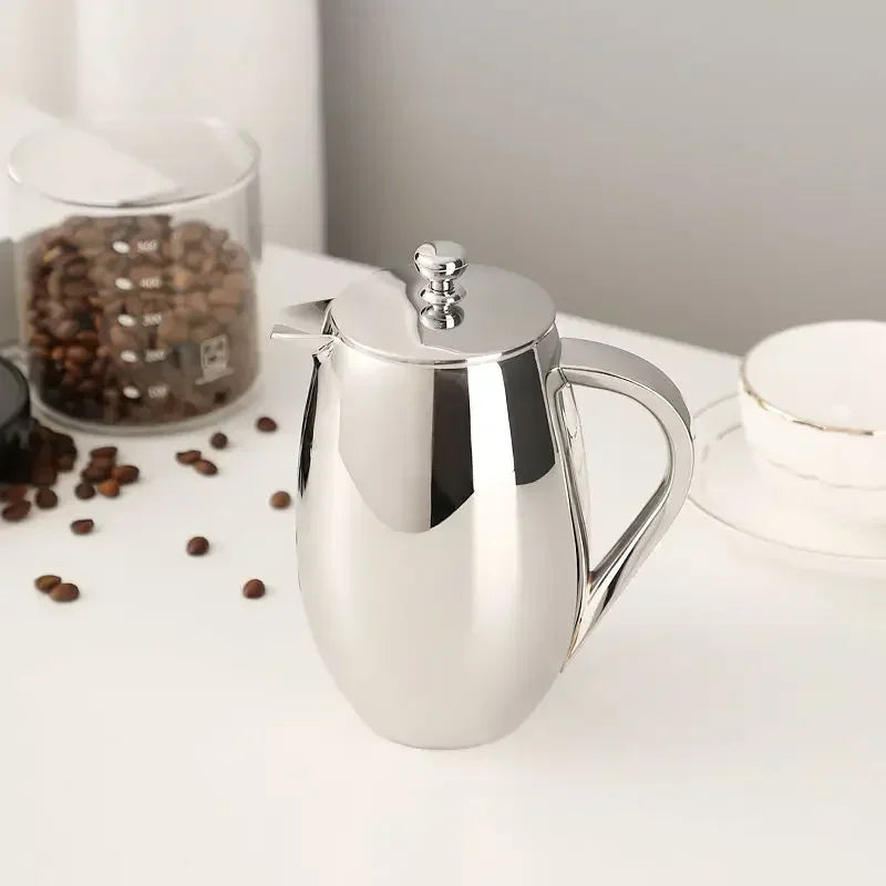 Stainless steel french press coffee pot on a white surface with coffee beans and a glass container in the background.