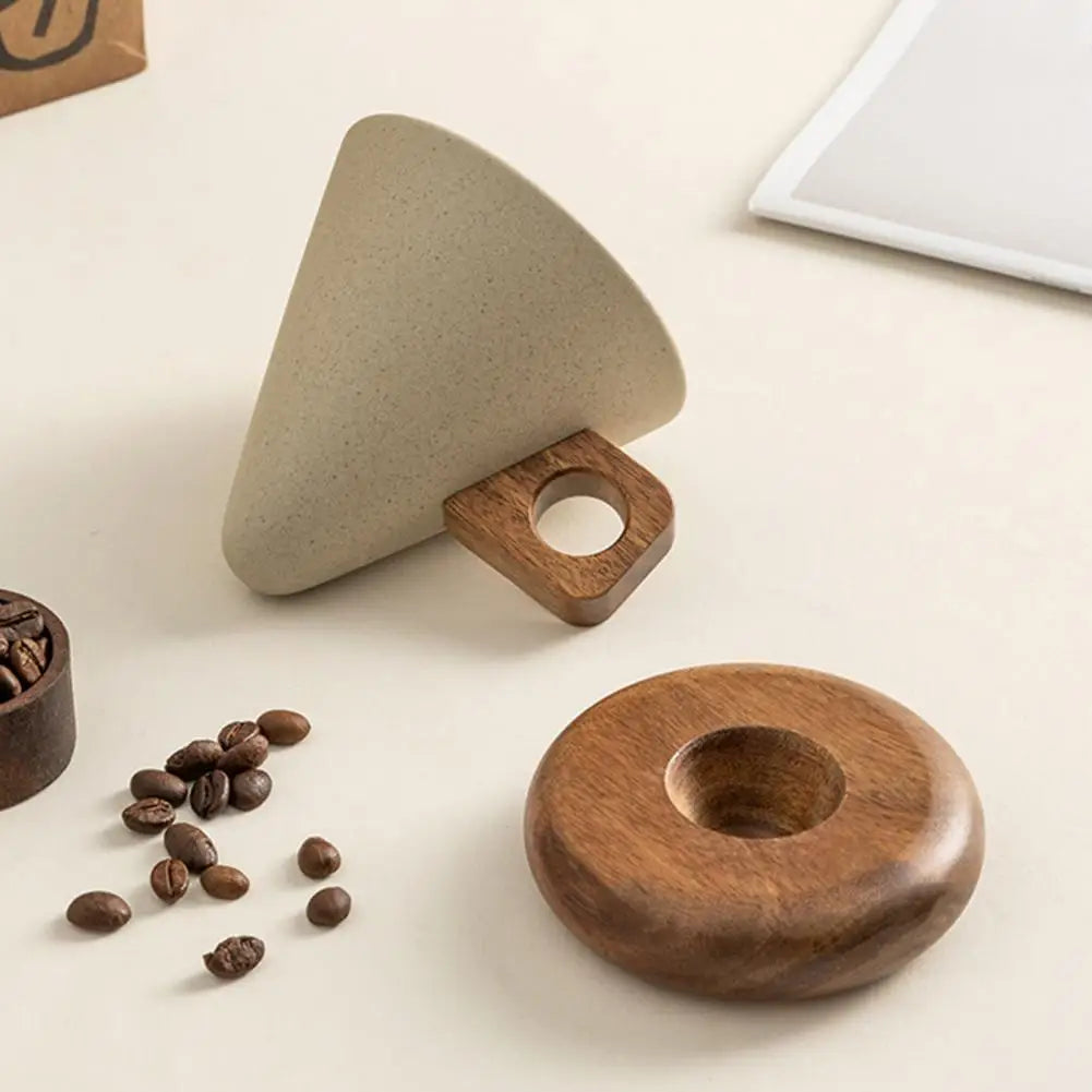 A minimalist coffee accessory set on a light beige surface, featuring a cone-shaped beige stone filter holder with a wooden handle, a round wooden coffee tray, and scattered coffee beans nearby.