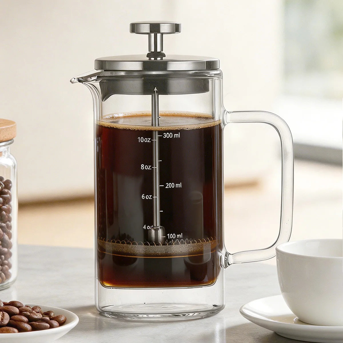 A clear glass French press coffee maker with a stainless steel lid and plunger, filled with dark coffee, shown on a countertop beside a white cup and a jar of coffee beans.