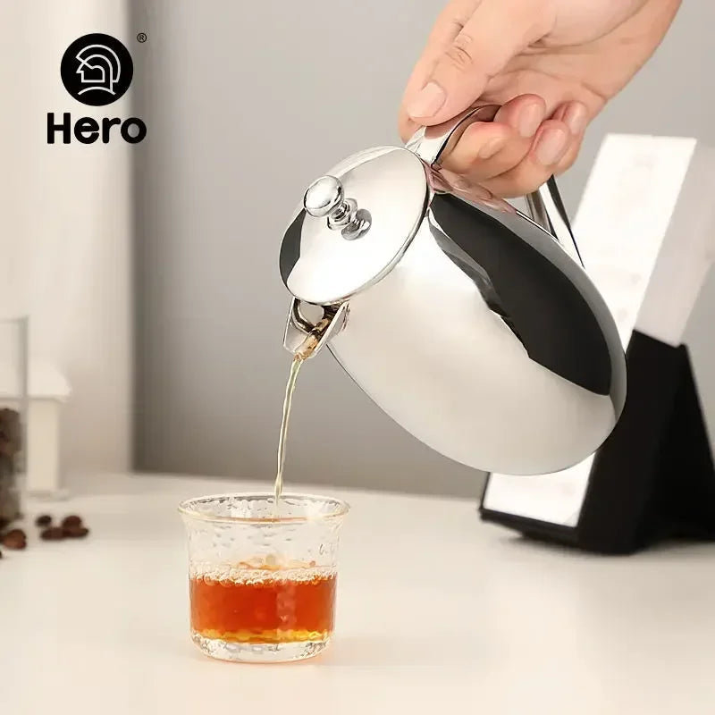Person pouring tea from a silver teapot into a glass cup on a white surface with 'Hero' branding.