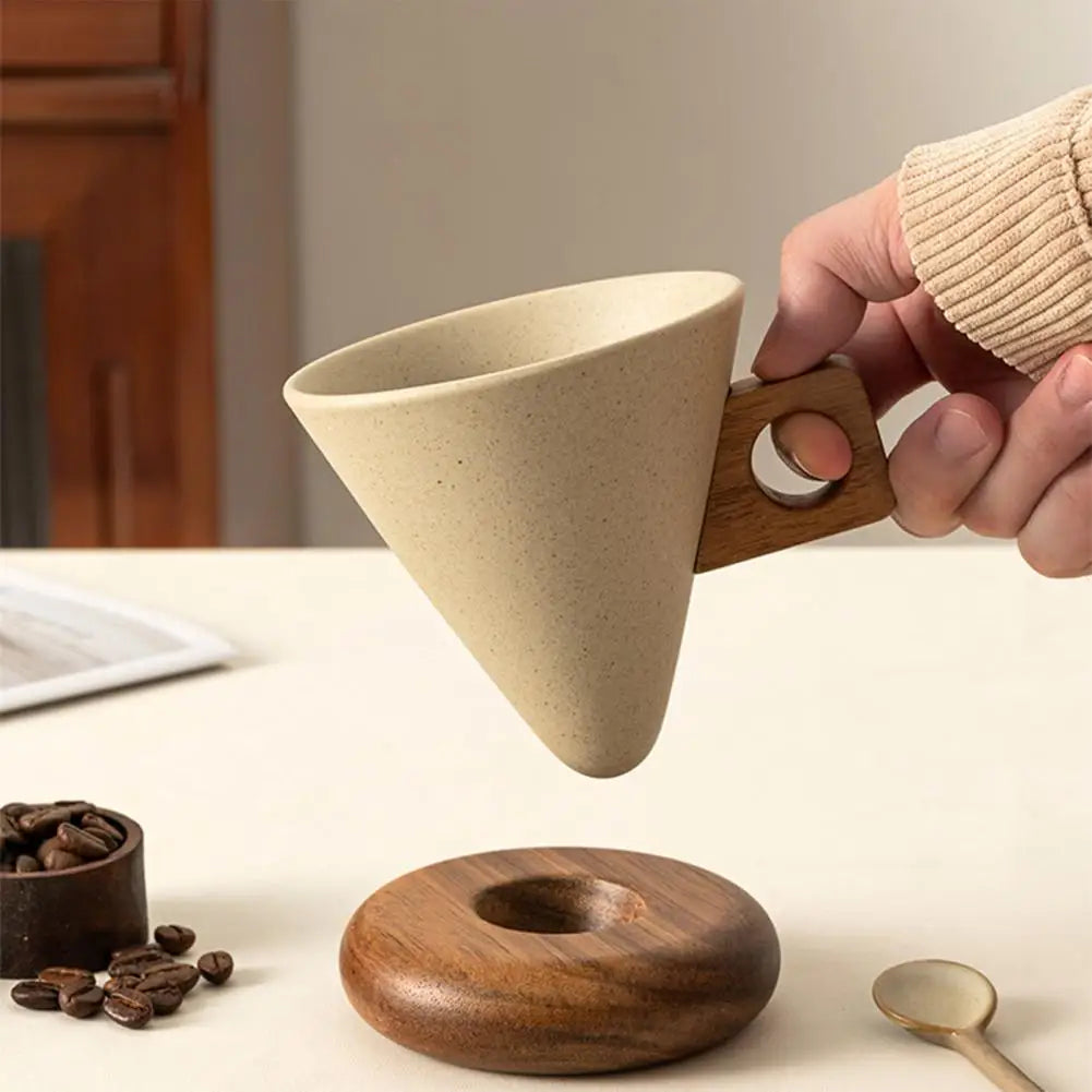 Hand holding a conical ceramic mug with wooden handle above its round wooden saucer, with coffee beans nearby