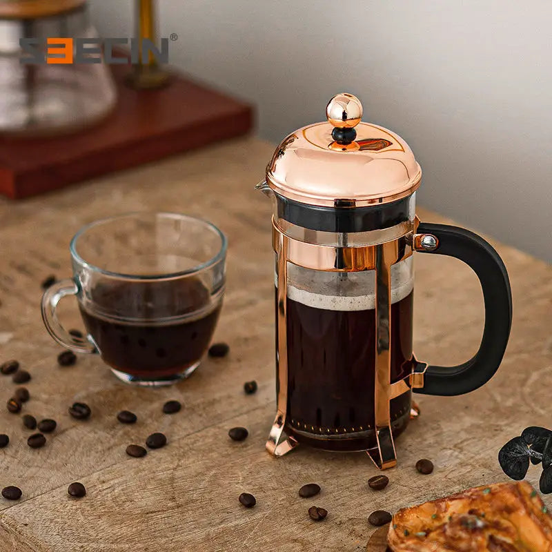 A copper and stainless steel French press with a black handle sits on a wooden table next to a glass cup of black coffee, with scattered coffee beans and a pastry in the foreground.
